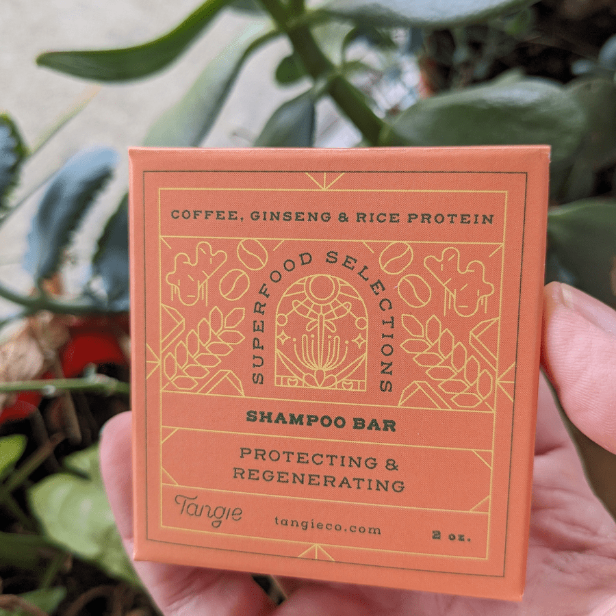 Person holding Tangie Superfoods Shampoo Bar against a lush green background, showcasing eco-friendly packaging and plant-based ingredients like caffeine, rice protein, and ginseng.